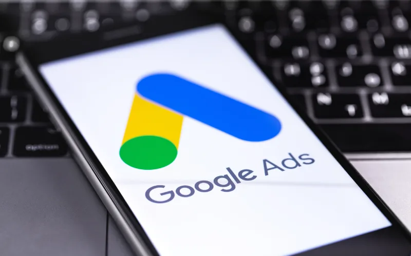 A smartphone displaying the Google Ads logo on a keyboard background. The logo features colorful geometric shapes.