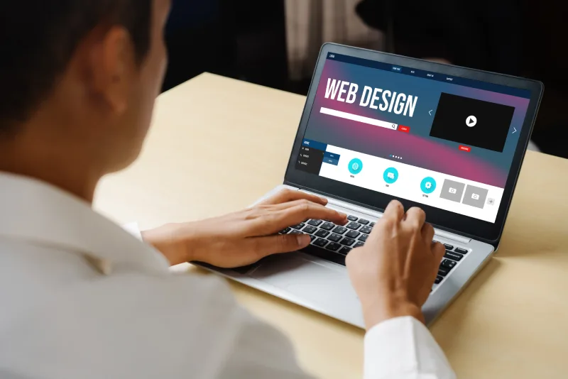 A person using a laptop on a desk, focused on a web design interface with video and editing tools displayed on the screen.